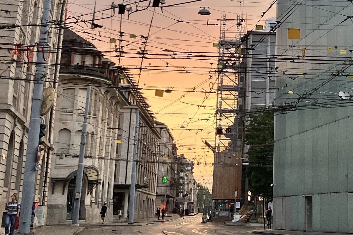 A city street at sunrise seen down its length, a dense canopy of tram wires crossing overhead and the sky turning orange between the buildings.