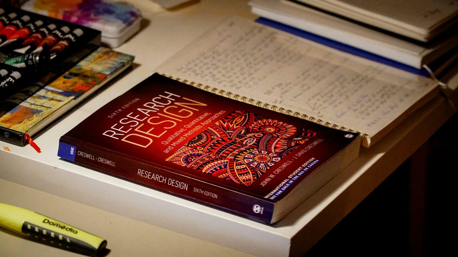 Creswell's Research Design lying on a desk beside an open notebook of handwritten notes, a highlighter and a paint box.