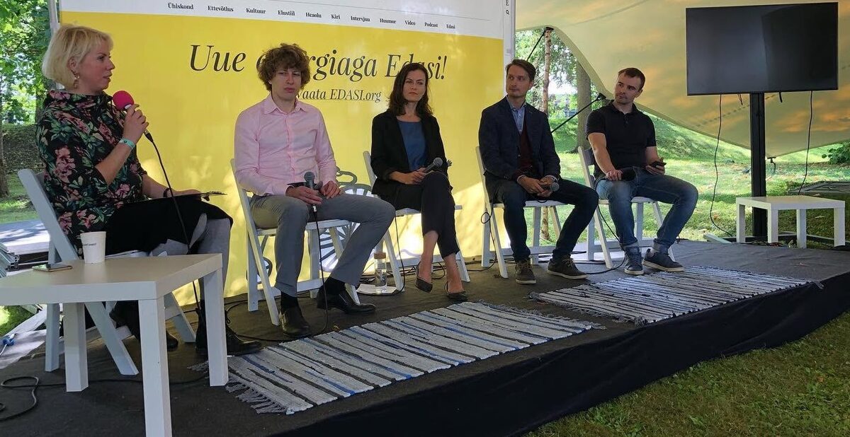 An outdoor panel under a white marquee: five people on stage in garden chairs, one speaking into a microphone.