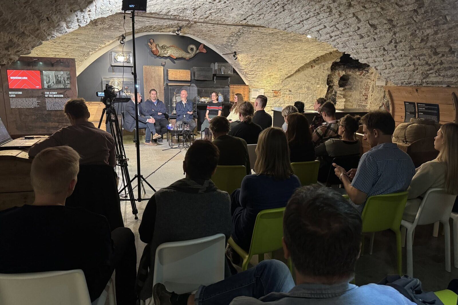 A vaulted stone cellar of a museum, seen from the back of a full audience, with five panellists seated under the arch.