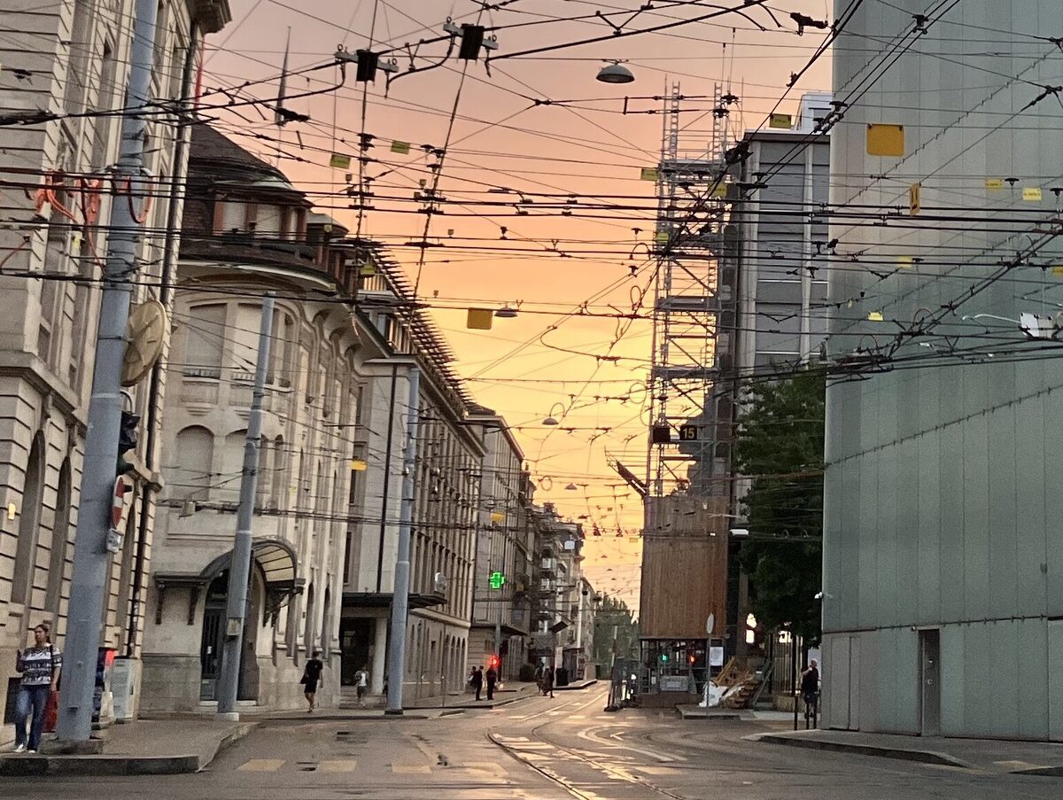 A city street at sunrise seen down its length, a dense canopy of tram wires crossing overhead and the sky turning orange between the buildings.